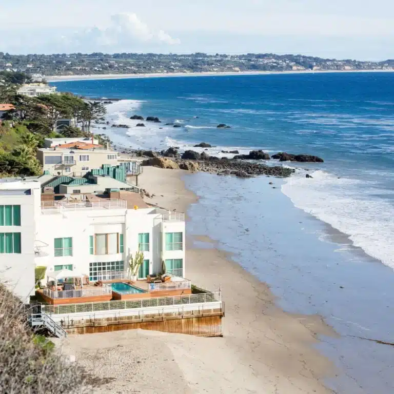 A scenic view of a luxury beachfront home with a private pool overlooking the ocean in Malibu, California, with waves gently reaching the sandy shore.