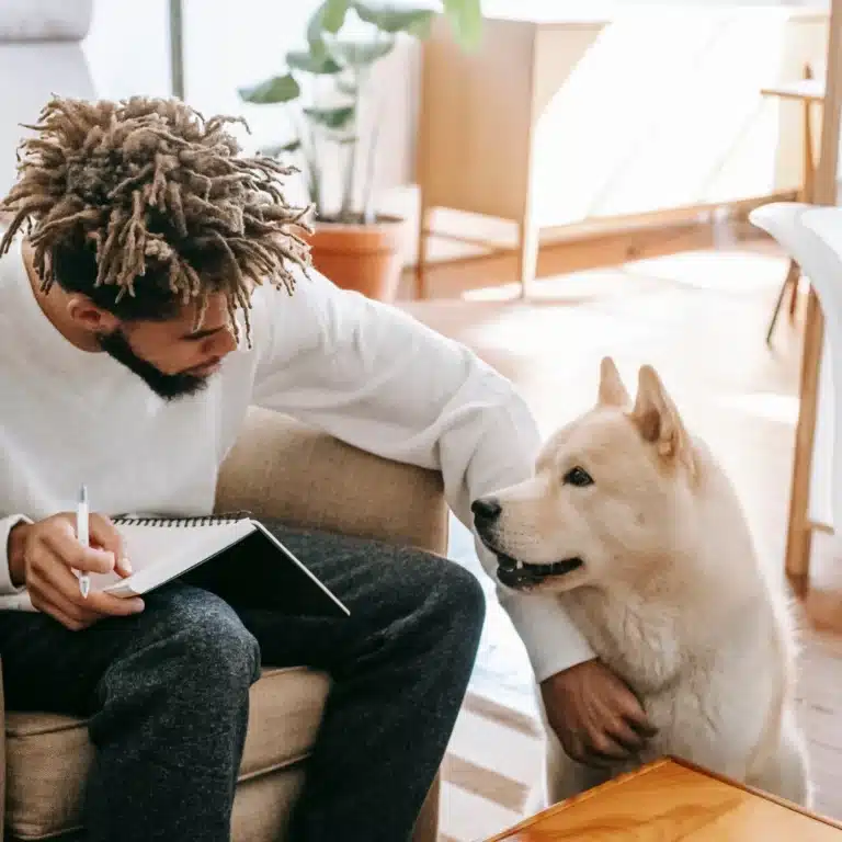 Man sitting in a cozy living room writing in a notebook while gently petting a friendly cream-colored dog.